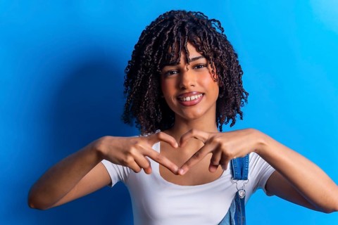 Smiling young woman making heart shape with hands, expressing love and affection on vibrant blue