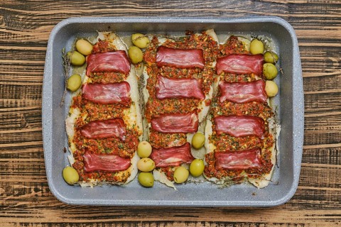 Roasted fish fillets are layered with bacon and herb sauce, accompanied by green olives. This