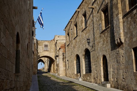 Historic street with stone buildings and arches, decorated with a Greek flag on a sunny day,