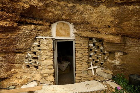 Entrance to a cave carved into the rock, surrounded by a stone wall, with a cross and plants on the