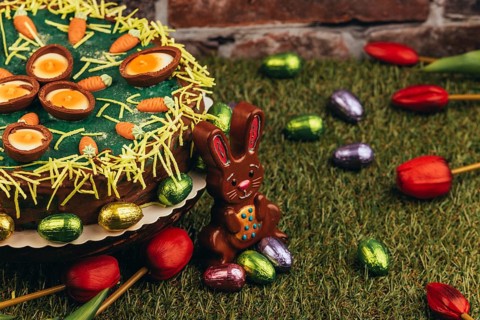 An Easter cake with carrots next to chocolate eggs, Easter bunnies and tulips on green grass