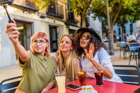 Three modern diverse women taking selfie with digital camera and tripod at the terrace of a bar