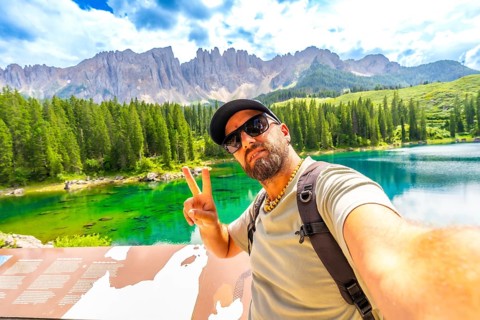 Tourist gesturing peace sign while taking a selfie at lake carezza with latemar mountain range in