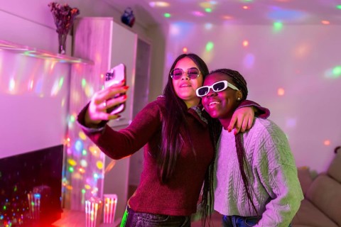 Two teenage girls wearing sunglasses are taking a selfie with a smartphone at a home party with