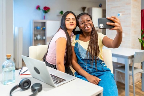 Two happy multi ethnic female students taking a selfie with a smartphone while studying at home