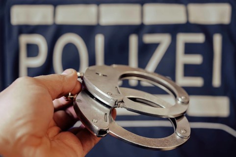 A pair of handcuffs is held by one hand, a police sign in the background