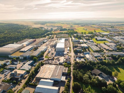 Numerous buildings and streets in an industrial area, surrounded by wooded landscape under a sunny