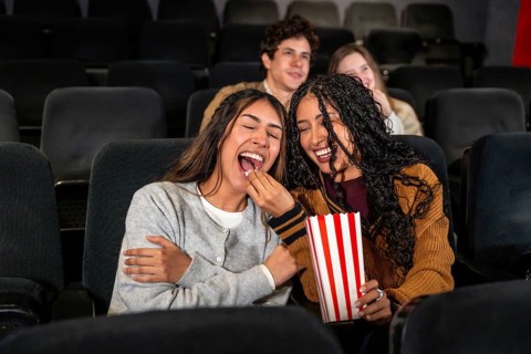Two young women are enjoying a movie at the cinema, laughing and sharing popcorn, with other people