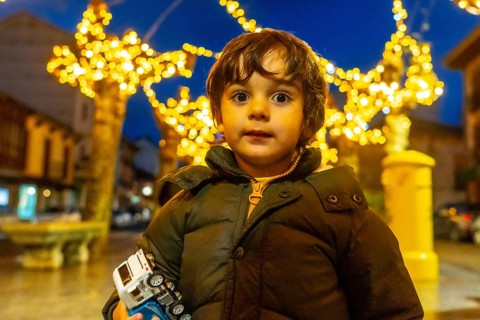 A young boy is standing in front of a Christmas tree with a toy car in his hand. The scene is