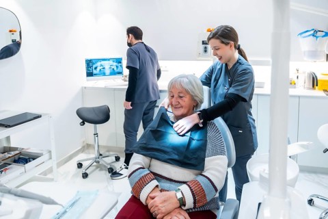 Dentist adjusting bib on senior woman for a dental checkup in a modern clinic, while a technician