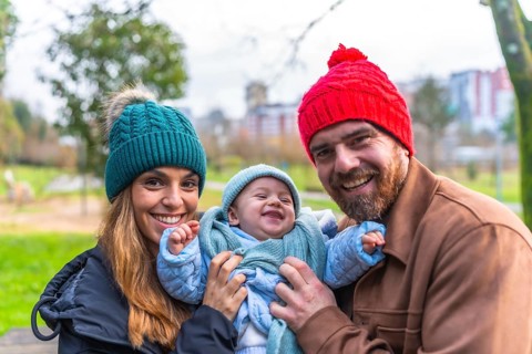 Smiling parents holding their laughing baby wearing winter clothes, enjoying a sunny winter day at