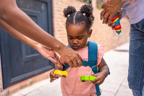 Baby african girl with schoolbag holding car toys standing in the street