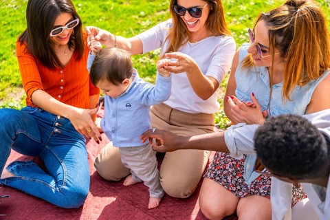Group of happy multi ethnic friends having fun with a baby learning to walk during a picnic in a