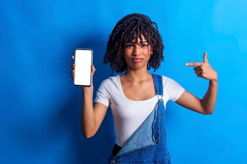Young woman showing a smartphone with blank white screen and pointing at it with index finger,