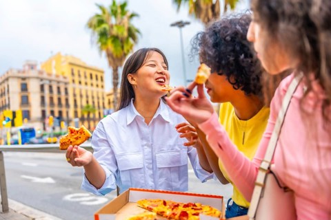 Three multiracial female friends chatting and eating pizza sitting on street