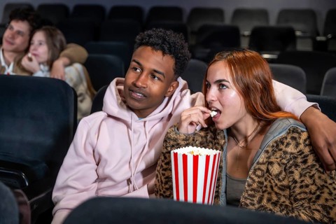 Young multi ethnic couple enjoying a movie and sharing popcorn in a cinema, surrounded by other