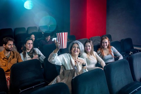 Mature woman enjoying a film in a movie theater, holding a popcorn bucket, surrounded by other