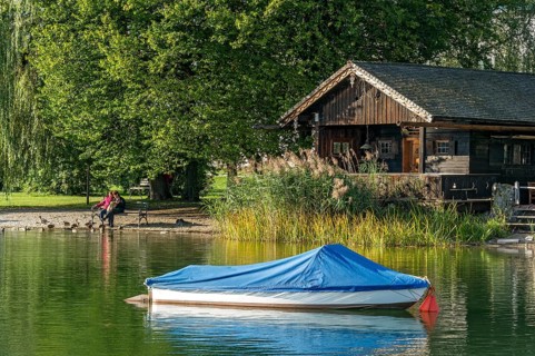 Wooden hut with common reed (Phragmites australis), covered boat, two woman on a park bench, shore