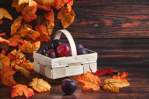 Plums in a wooden basket surrounded by orange autumn leaves