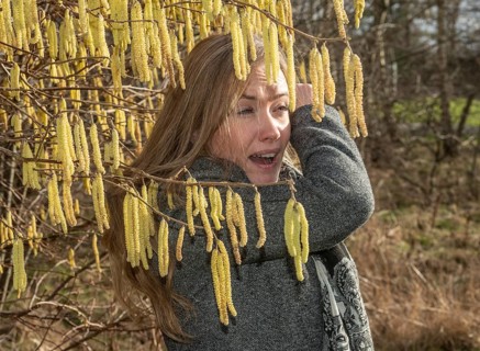 Young Caucasian woman with allergy problem stands by flowering hazel bush and sneezes in the crook