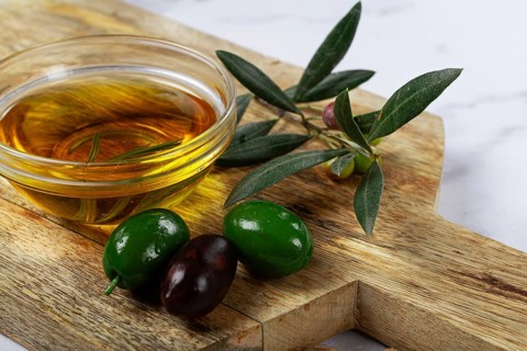A glass bowl filled with rich olive oil sits on a wooden cutting board alongside fresh green and