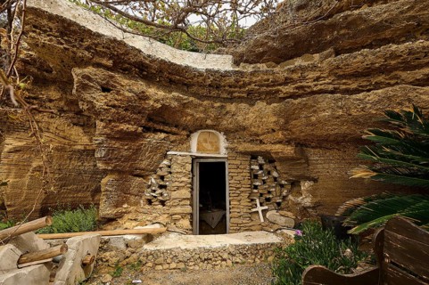 Exterior view of a cave carved into the rock with a stone wall and vegetation around the entrance,