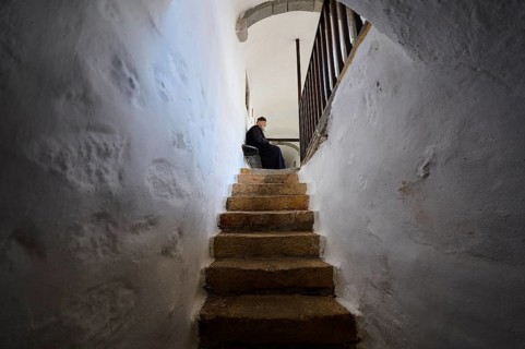 A monk sits on a bench at the end of a stone staircase in a narrow corridor of the monastery,