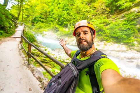 Tourist wearing an orange helmet and backpack takes a selfie while visiting vintgar gorge near bled