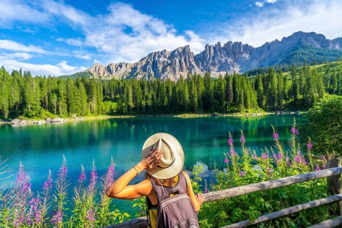 Woman with backpack and hat admiring the turquoise waters of lake carezza with the latemar mountain