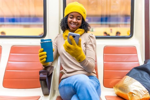 Smiling black woman wearing yellow winter clothes using phone and drinking coffee while traveling