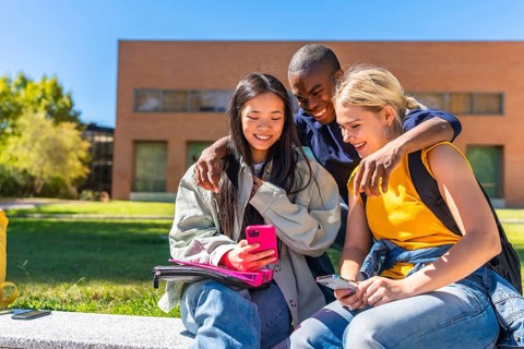 Happy multi-ethnic students using mobile and sitting outside the university campus in a sunny day