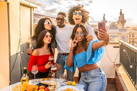 Happy multi-ethnic friends taking selfie celebrating evening party in rooftop drinking wine and