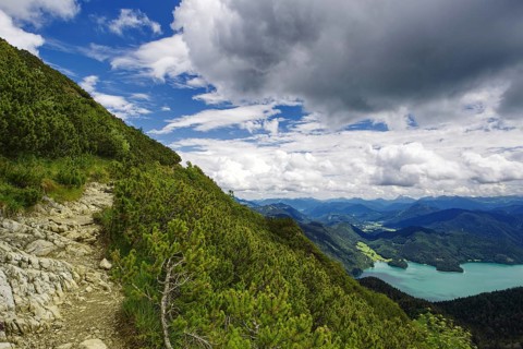 A stony hiking trail along a wooded mountain slope with sweeping views down into the valley to Lake