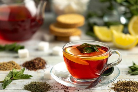 A cup of red tea with lemon and mint surrounded by sugar cubes, teas and biscuits