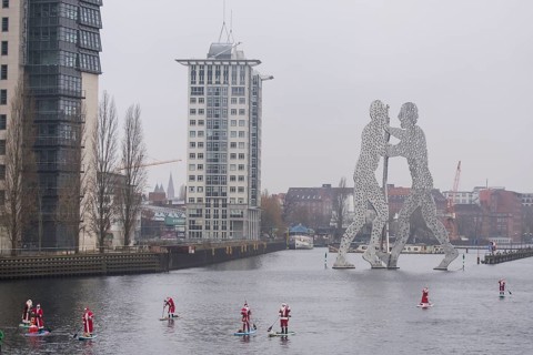 Water sports enthusiasts dressed as Santas ride SUPs on the Spree in front of the Molecule Man in