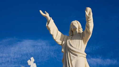 A stone statue of Jesus with outstretched arms against a blue sky, Agios Nikolaos church, cemetery,