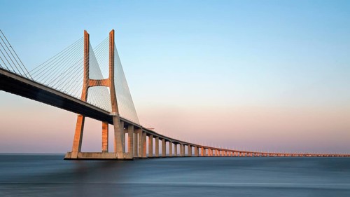 A long cable-stayed bridge spans over water under a gradient sunset sky, Lissabon, Vasco Da