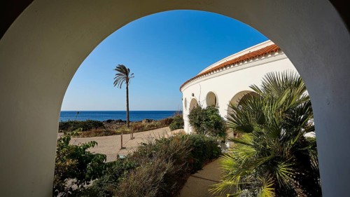 View through an arch of a circular building to the sea, surrounded by green plants and bushes,