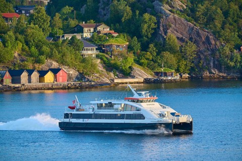 Colourful wooden houses by the water, surrounded by dense forest and hills, a ferry sails on the