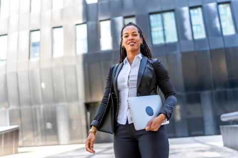 Professional young latin businesswoman standing carrying laptop in the city next to a modern