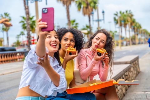 Multiracial female friends eating pizza and taking a selfie sitting in the street