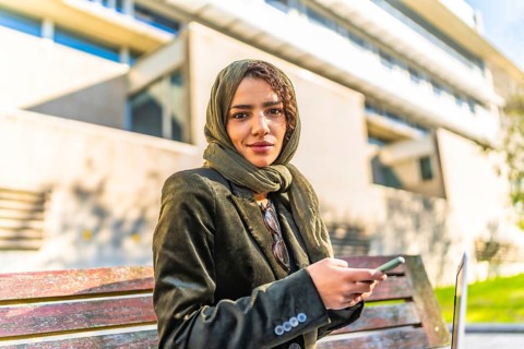 Muslim businesswoman using smartphone sitting on a park bench next to office building