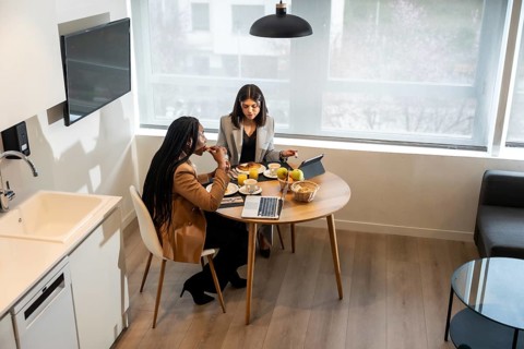 Two businesswomen are having breakfast in their hotel room while working on a tablet and laptop