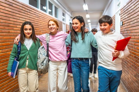 Four cheerful students are walking together in school corridor, carrying backpacks and notebooks