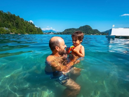 Father playing with his son in the clear water of lake bled, with mountains and forest in the
