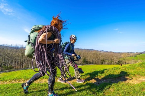 Two female climbers are walking across a mountain meadow carrying climbing rope and gear on a sunny
