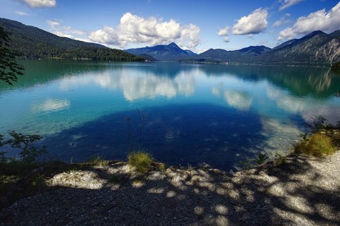A calm lake reflecting the clouds and mountains, framed by wooded hills, Walchensee, Kochel am See,