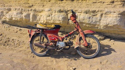 An old Honda C50 motorbike stands abandoned in front of a rocky cliff, Paralia Agios Theodoros