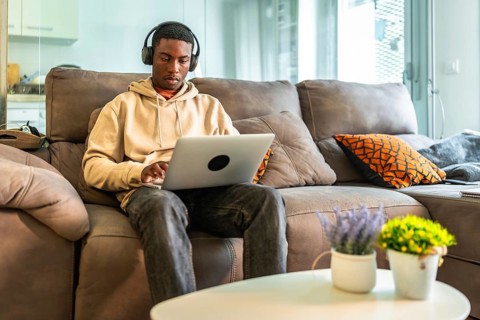 Young man concentrating on his laptop while wearing headphones, comfortably seated on a sofa in his