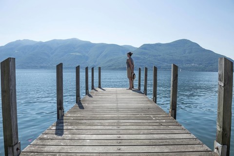 Woman Standing on a Pier on Lake Maggiore with Mountain in a Sunny Day in Brissago, Ticino,